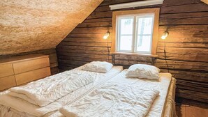 Quarto com teto inclinado de madeira, cama de casal com roupa de cama branca e duas luminárias fixadas na parede.