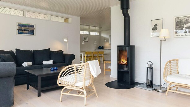 A modern living room with a black fireplace, a grey sofa, wicker chairs, and a dining area in the background. - 6 Person Holiday Home in Gilleleje (Gilleleje)