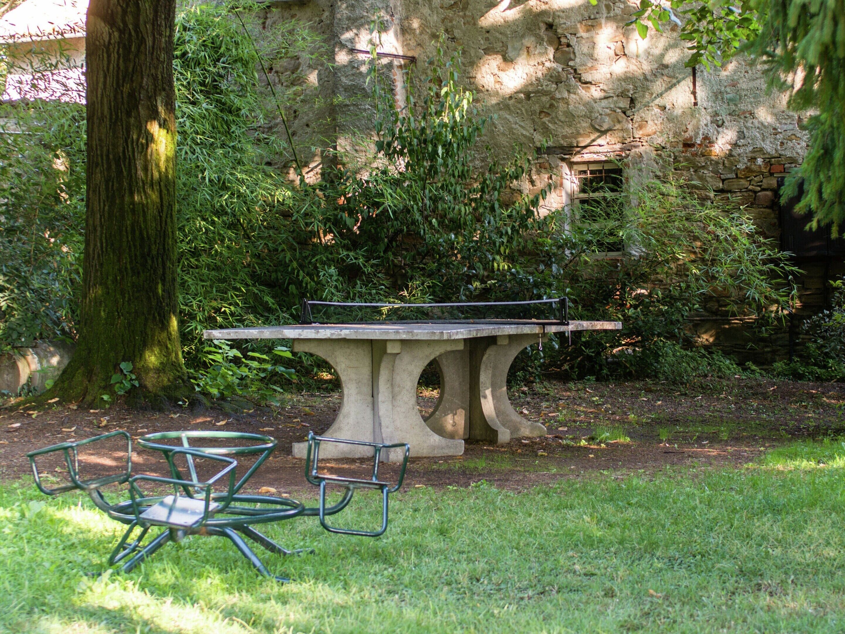 Una mesa de piedra con sillas en un jardín, con un árbol y un muro de piedra al fondo.
