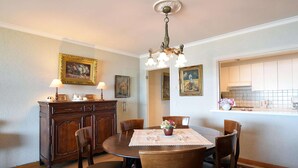 Un coin repas avec une table ronde en bois, six chaises, un lustre, un buffet avec un miroir, et une cuisine visible à travers une porte ouverte.
