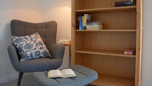 A grey armchair with a floral pillow and a small ottoman with an open book. A wooden bookshelf with books and a lamp is in the background. - Pleasant Apartment in Stadtilm (Stadtilm)