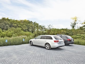 A parking area with two cars, a white sedan and a black sedan, parked on a paved surface with a grassy area and trees in the background. - Holiday Home sea Ambience (Karlshagen)