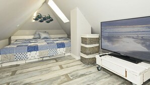 Ein Schlafzimmer mit schrägem Dach, einem Bett mit gemustertem Überwurf, einem Fernseher auf einem weißen Wagen und Blick auf den Strand durch das Fenster.