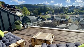Una terraza en la azotea con mobiliario de madera, una planta en maceta y vista a un pueblo con casas y un puente al fondo.