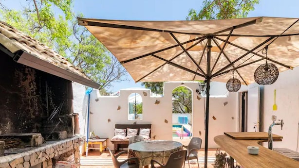 A patio area with a stone fireplace, a round table with chairs, and a large umbrella providing shade. - Belvilla by OYO Casa Tata (Granadilla de Abona)