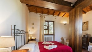 A bedroom with a wooden ceiling, a bed with a red bedspread, a nightstand with a lamp, and a window with curtains. - Villa Montecchio Comfortable Holiday Residence (Cortona)