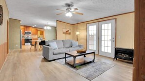 A spacious living room with a grey sofa, a coffee table, and a fireplace. A kitchen is visible in the background with wooden cabinets and a microwave.