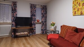 A living room with a TV on a wooden stand, a red sofa, a small table with a vase of flowers, and curtains with floral patterns.