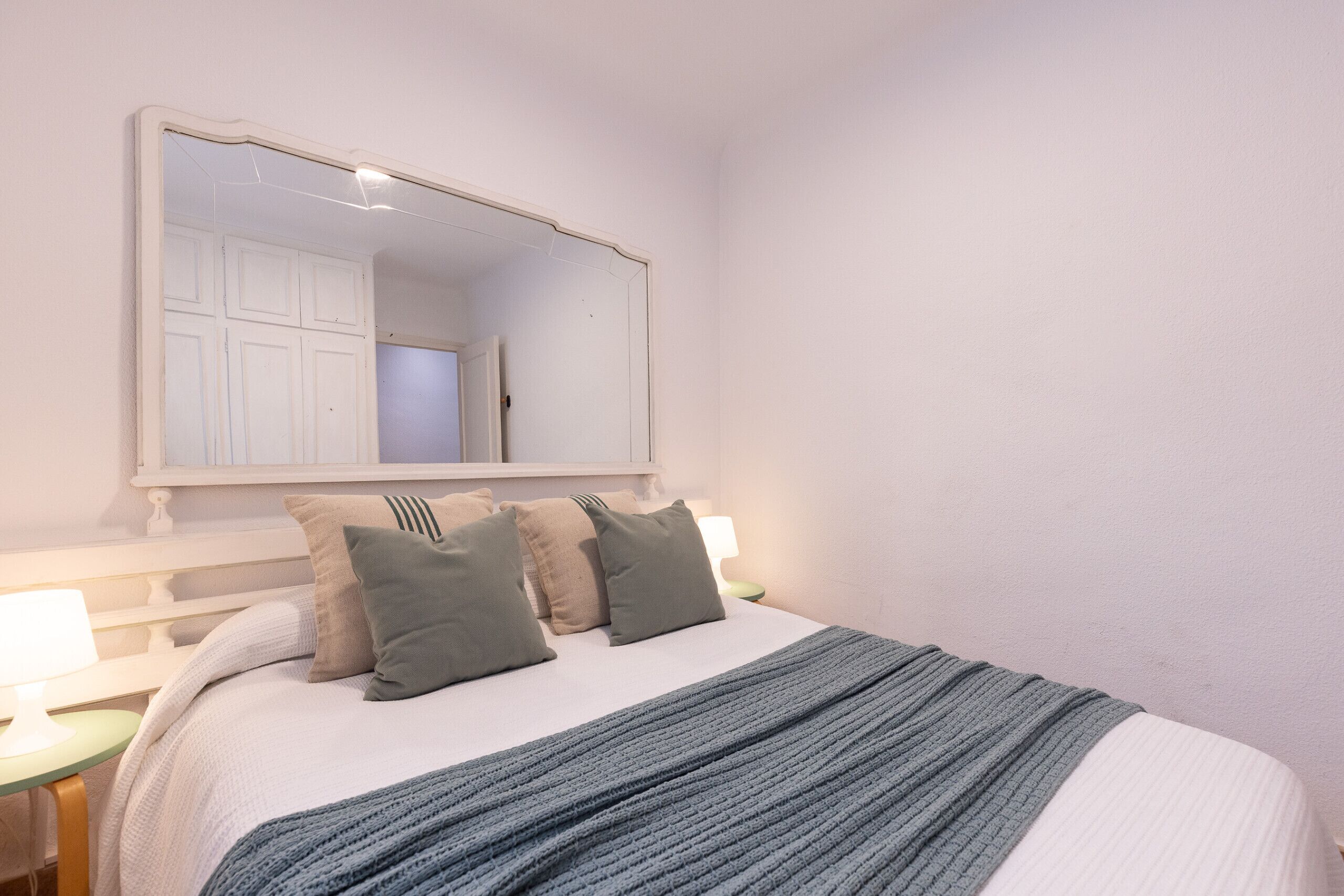 A bedroom with a large mirror, a bed with a grey blanket, and two bedside tables with lamps.