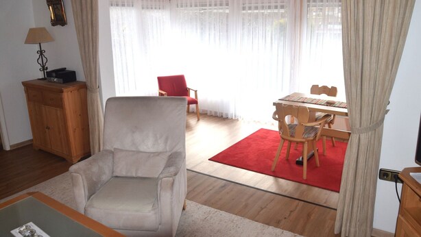 A living room with a beige armchair, a wooden cabinet, a dining table with chairs, and a red rug. - Ferienhaus im Malerischen Allgau (Fuessen)