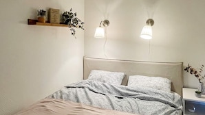 A bedroom with a bed, bedside table, wall-mounted lights, and a shelf with plants.