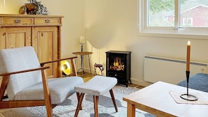 A cozy living room with a fireplace, a wooden cabinet, a chair, a table, and a window with a view of trees and a house.