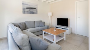 Un salon moderne avec un canapé d’angle gris, une table basse en bois, un téléviseur à écran plat posé sur un meuble blanc et un plafond avec des poutres apparentes.