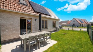 Un immeuble résidentiel doté d’un toit en tuiles, de panneaux solaires, d’une façade en briques et d’un patio aménagé avec du mobilier d’extérieur.