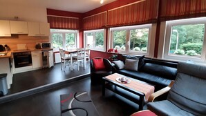 Una cocina moderna con armarios blancos, un comedor con sofá negro y sillas rojas, y ventanas amplias que dan a zonas verdes.