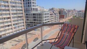 Un balcón con una silla a rayas y una mesita pequeña, con vista a un paisaje urbano que incluye edificios modernos y una calle con coches.