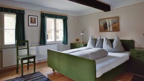 A bedroom with a green bed, a chair, a radiator, a window with curtains, a nightstand, and a painting on the wall. - The Holler Comfortable Holiday Residence (Schwanberg)