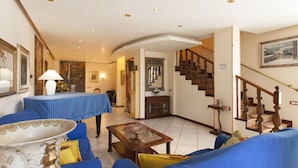 A living room with a blue sofa, a coffee table, and a table with a blue tablecloth. There is a staircase with wooden railings leading to another room. - Topazio Comfortable Holiday Residence (Quartu Sant'Elena)