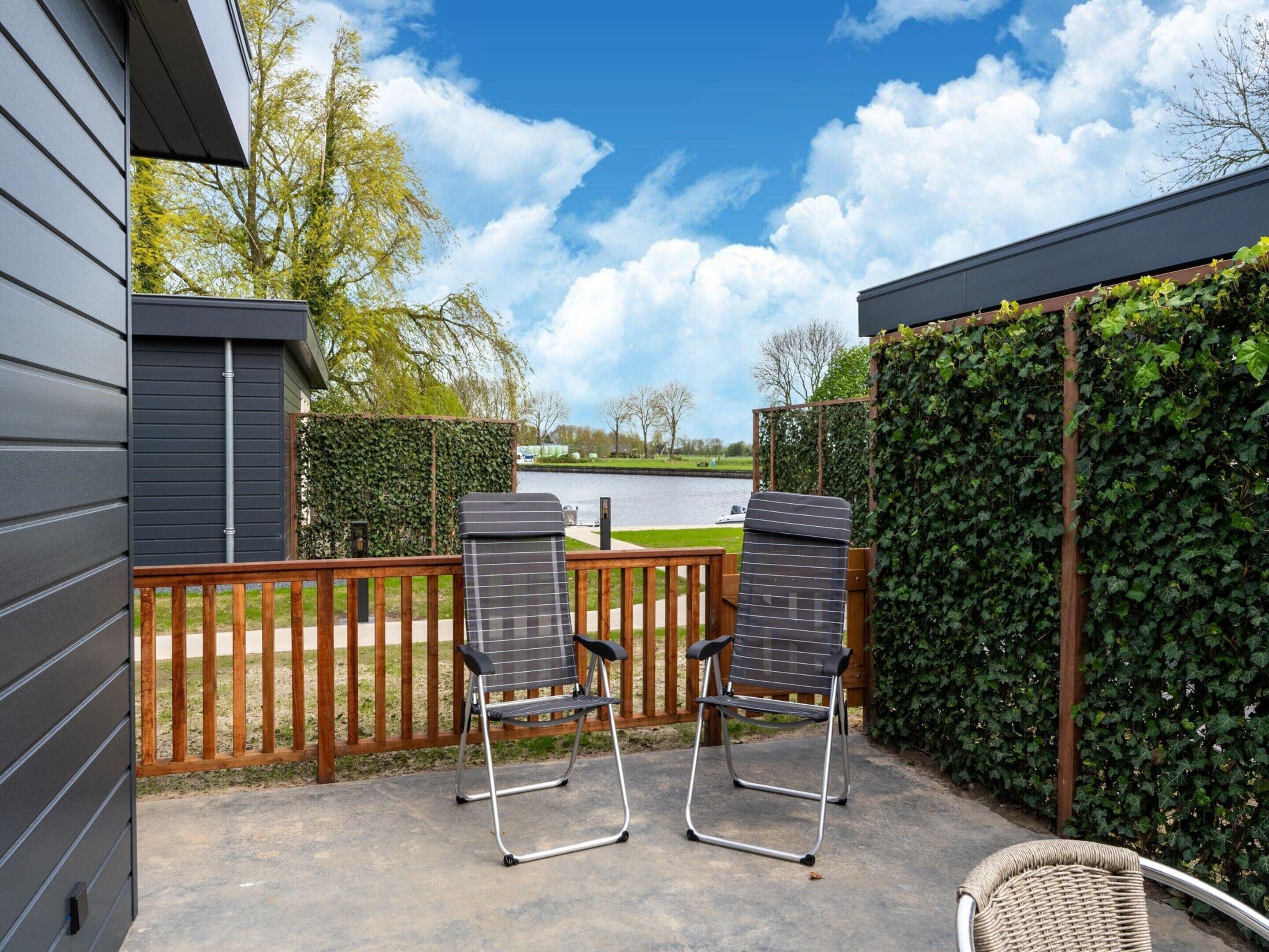 A patio with two metal chairs and a wicker chair, a wooden fence, and a view of a river and trees.