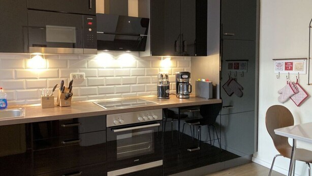 A modern kitchen with black cabinets, a stainless steel range hood, a built-in oven, a coffee maker, and a small dining area with a chair. - Stadthaus Emmy Marxen (Schleswig)