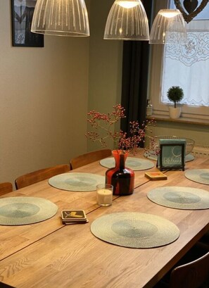 A wooden dining table with placemats, a candle, a vase with branches, and a framed picture. - Stadthaus Emmy Marxen (Schleswig)