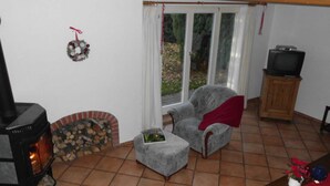 Un salon chaleureux avec une cheminée, un fauteuil gris, une petite table recouverte d’une nappe rouge et un téléviseur posé sur un meuble en bois.