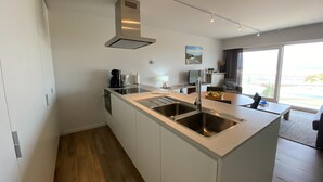 A modern kitchen with a central island, stainless steel appliances, and a large window offering a view. - Apartment in Nieuwpoort With sea View (Nieuwpoort)