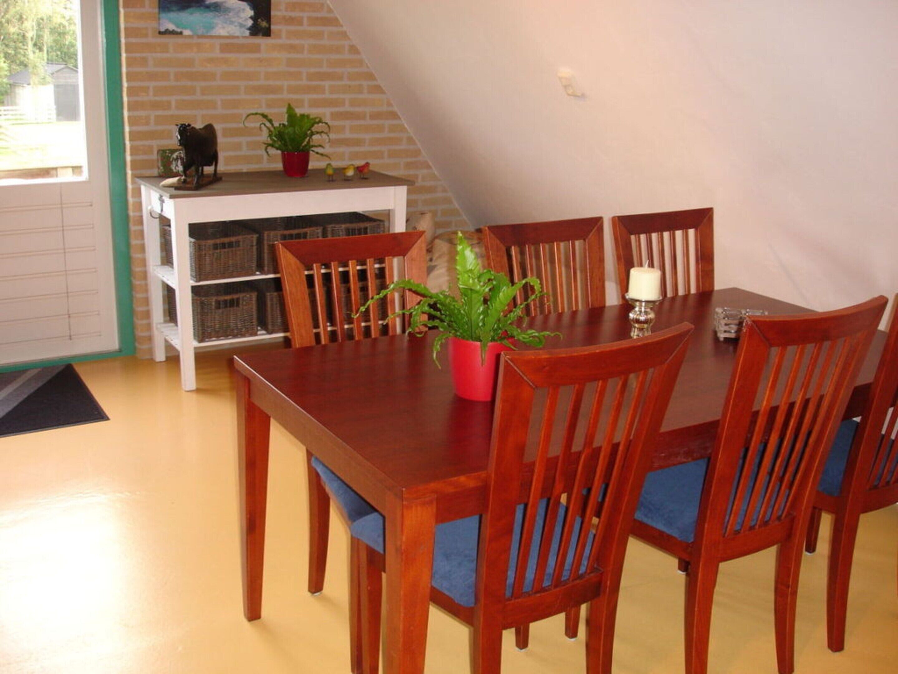 A dining area with a wooden table and chairs, a potted plant, and a small shelf with decorative items.