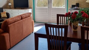 A cozy living room with a brown sofa, a wooden dining table with chairs, a TV on a cabinet, and a door with a view of trees. - Country Life Comfortable Holiday Residence (t Zand)