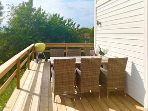 A wooden deck with a table and wicker chairs, a grill, and a white building in the background. - 5 Star Holiday Home in Borgholm (Borgholm)