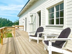 A wooden deck with outdoor furniture, including two chairs and a table, in front of a white building with large windows. - 5 Star Holiday Home in Borgholm (Borgholm)