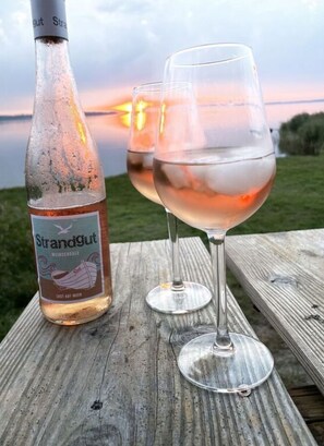 Une bouteille de vin Strandgut et deux verres sur une table en bois, avec un coucher de soleil en arrière-plan.