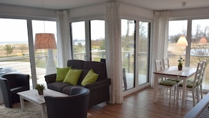 A modern living room with a sofa, chairs, a dining table, and large windows offering a view of the outdoors. - Dune View No. 3 Comfortable Vacation Home (Heiligenhafen)