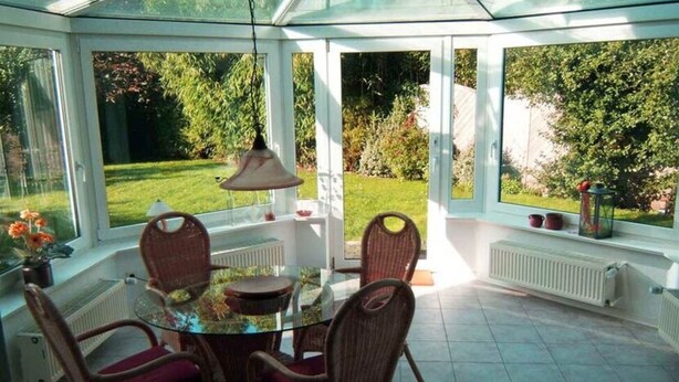 A conservatory with a glass table surrounded by wicker chairs, a hanging lamp, and a view of a garden. - Heinrich Tiarksstrasse 36 (Wangerland)