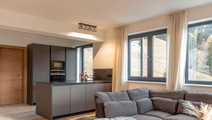 A modern kitchen with grey cabinets and a built-in microwave, a grey sofa with a throw blanket, and large windows with curtains. - Apartment With Infinity Pool (Sankt Martin am Tennengebirge)