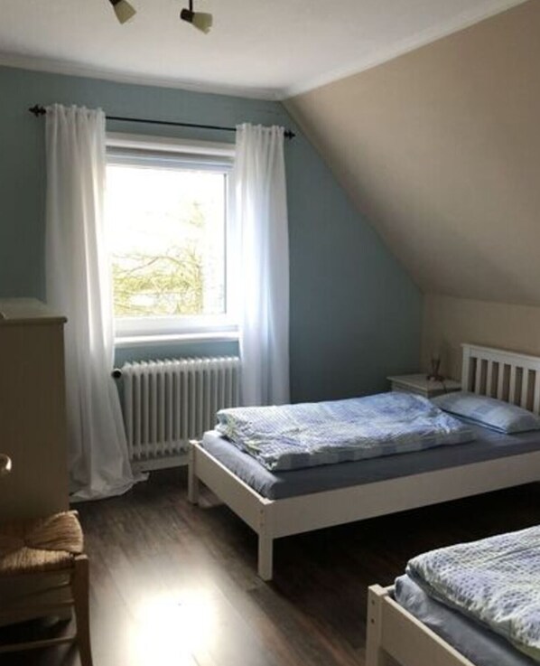 Une chambre avec un plafond en pente, un lit avec un cadre blanc, une table de chevet, un radiateur et une fenêtre avec des rideaux blancs.