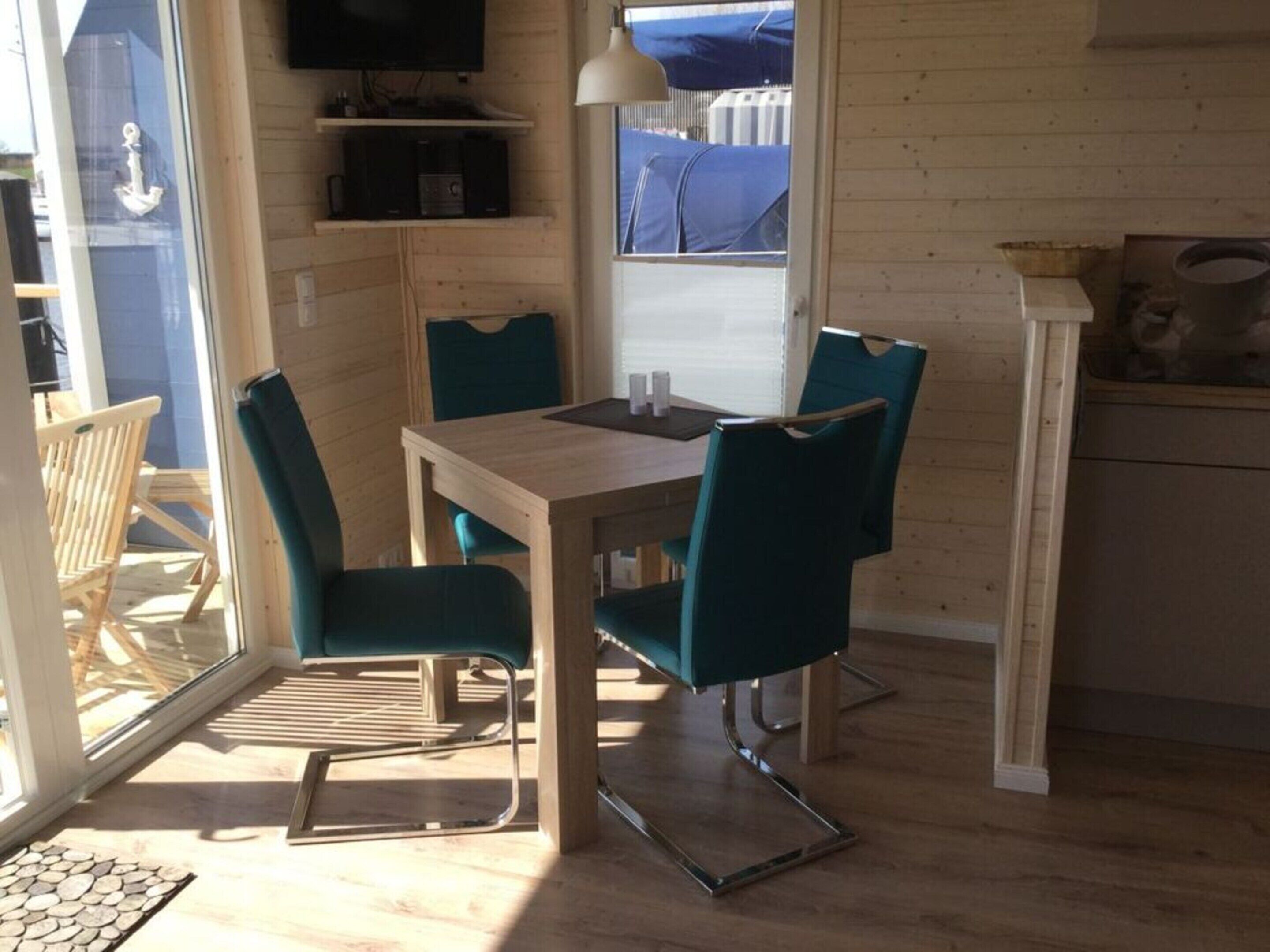 A dining area with a wooden table and four teal chairs, a television above the table, and a view of the outside through a window.