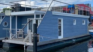 Una casa flotante azul atracada en un puerto deportivo, con una pequeña terraza y zona de estar al aire libre.