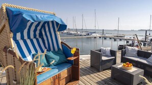 Una terraza con mobiliario de mimbre, incluyendo una silla azul y blanca a rayas y un sofá de mimbre, con vista al puerto deportivo y barcos al fondo.