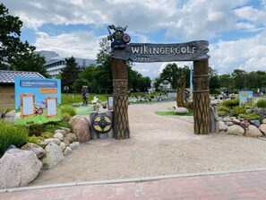 Uma placa de madeira com a inscrição 'WIKINGERGOLF OAMP', rodeada por pedras e vegetação.