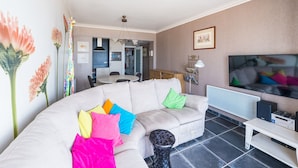 A living room with a beige sofa, colorful cushions, a TV, and a dining area with a table and chairs. - Apartment on the 7th Floor With sea View (Middelkerke)
