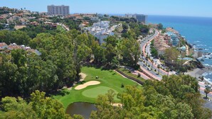 Un campo de golf con un estanque, rodeado de árboles densos y una carretera costera con edificios.