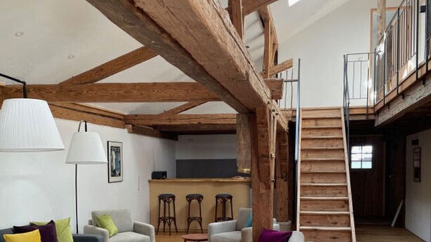 Un loft spacieux avec des poutres apparentes, une mezzanine et un espace de vie moderne à aire ouverte, comprenant des canapés et une table basse.