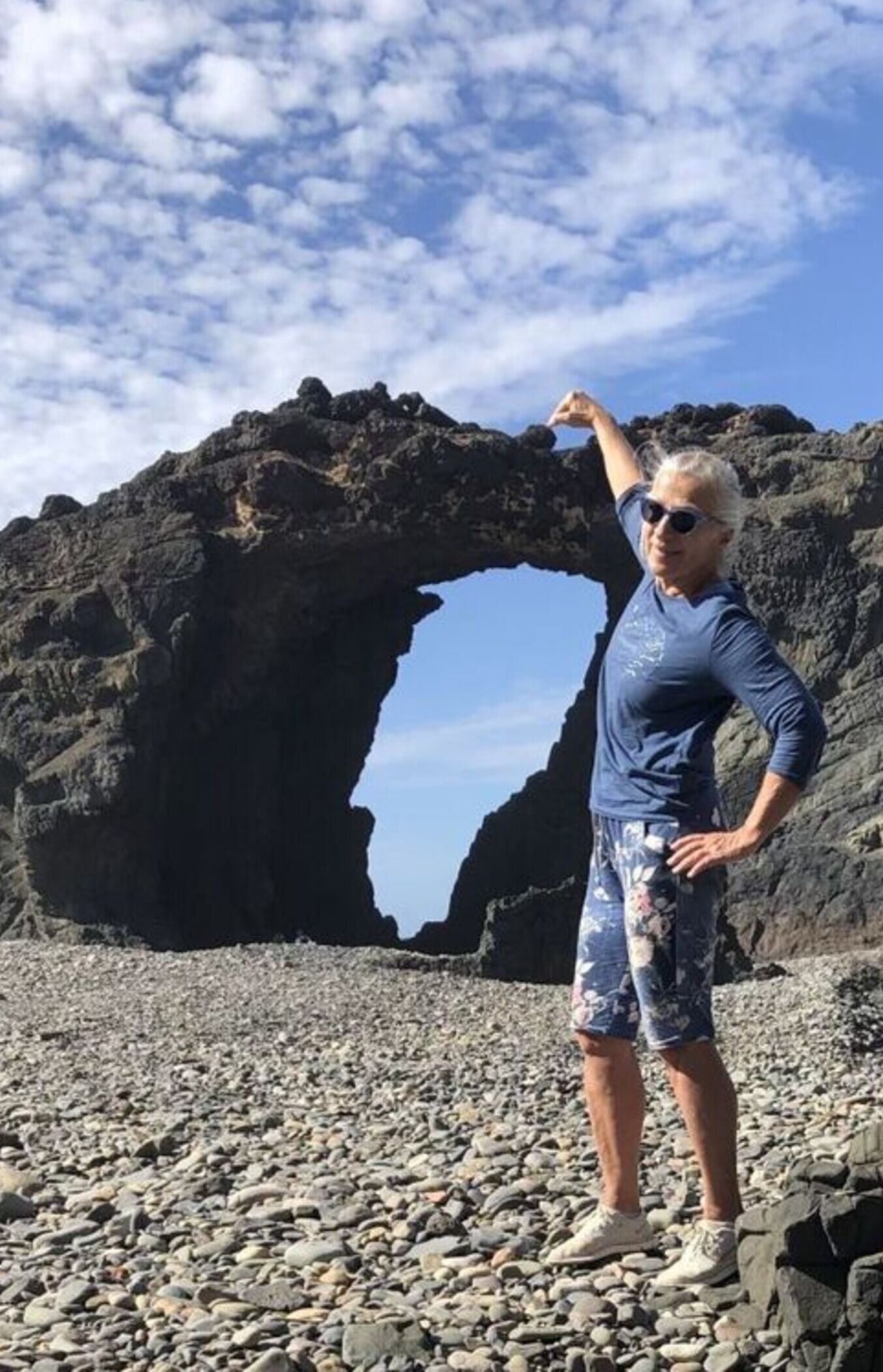 Une personne se tient sur une plage rocheuse, avec une grande arche rocheuse en arrière-plan.