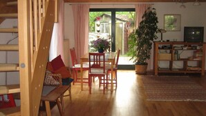 A cozy room with a wooden staircase, a dining area with chairs and a table, a bookshelf, a television, and a large potted plant. - Nice Apartment in Lubmin (Lubmin)
