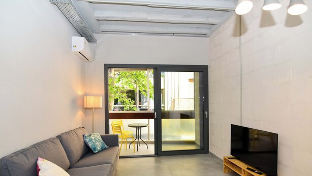 Uma sala de estar moderna com um sofá cinzento, uma televisão de tela plana em um suporte de madeira e uma porta de vidro deslizante que dá para uma varanda externa com cadeiras amarelas e uma mesa.