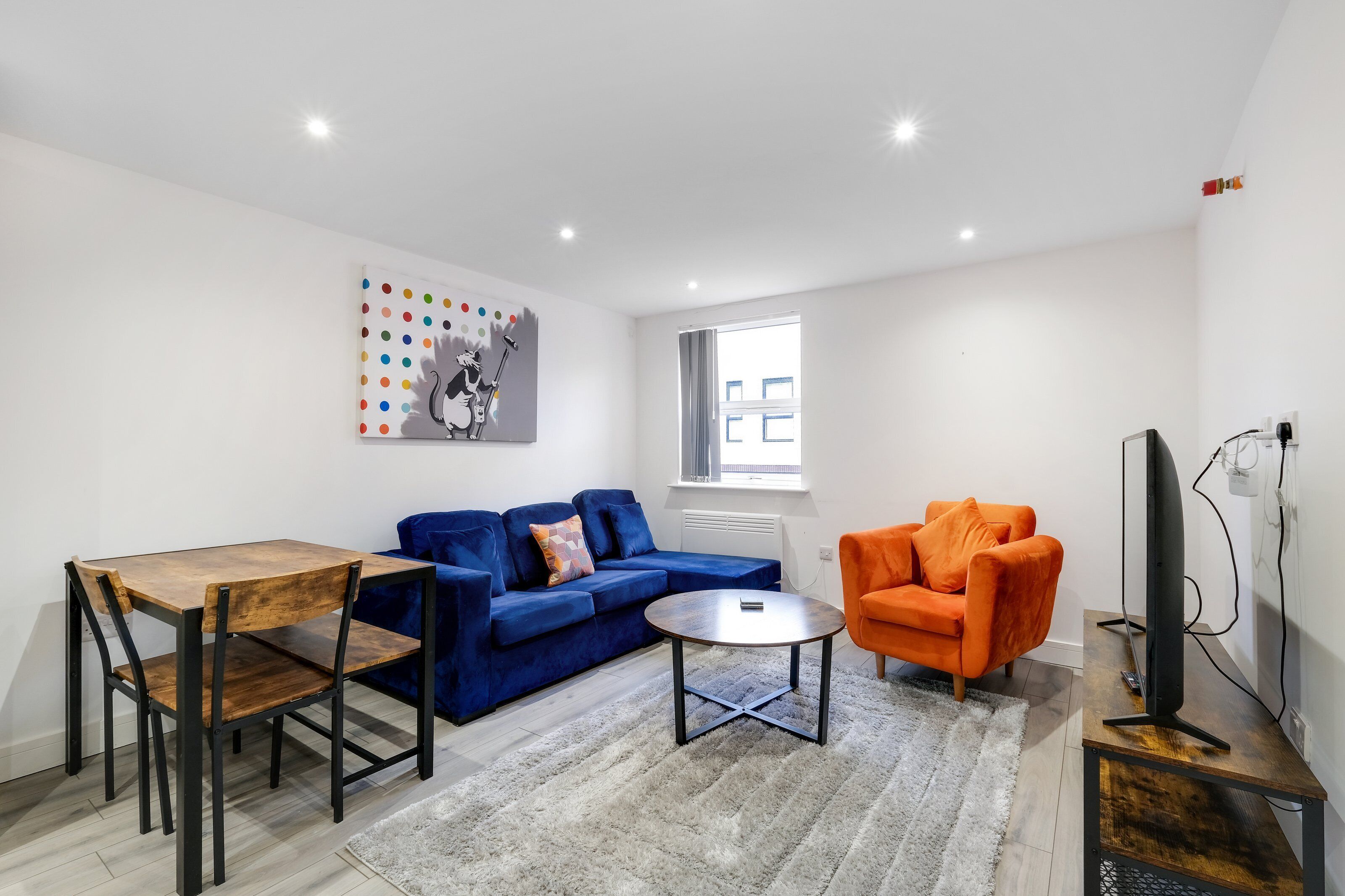 A modern living room with a blue sofa, an orange armchair, a wooden dining table, and a TV on a wooden stand.