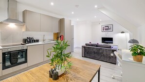 A modern kitchen with built-in appliances, a dining area with a wooden table, and a living space with a sofa and TV. - Modern 1 Bedroom Apartment - Camberley (Camberley)