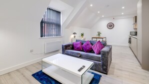 A modern apartment with a loft-style ceiling, a grey sofa with purple cushions, a white coffee table, a small side table with a lamp, and a kitchen area. - Modern 1 Bedroom Apartment - Camberley (Camberley)
