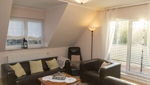 Uma sala de estar acolhedora com um sofá, poltronas, uma mesa de centro e uma luminária. Há uma janela com cortinas e um radiador na parede.
